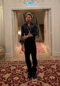 Jeune homme en tenue noire avec un grand collier croix, debout dans un couloir orné avec un tapis à motifs et un escalier derrière lui.