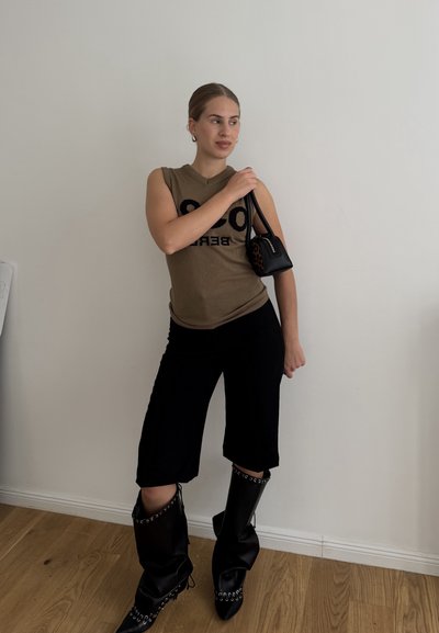 Top sin mangas de color bronce, con un estampado negro audaz, pantalones cortos negros y botas altas negras con detalles de hardware plateado. Pequeño bolso negro con acentos.