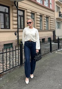 Femme portant un pull blanc boutonné, jean foncé, talons blancs, lunettes de soleil noires, tenant un sac à main noir sur un trottoir en ville près d'un bâtiment beige.