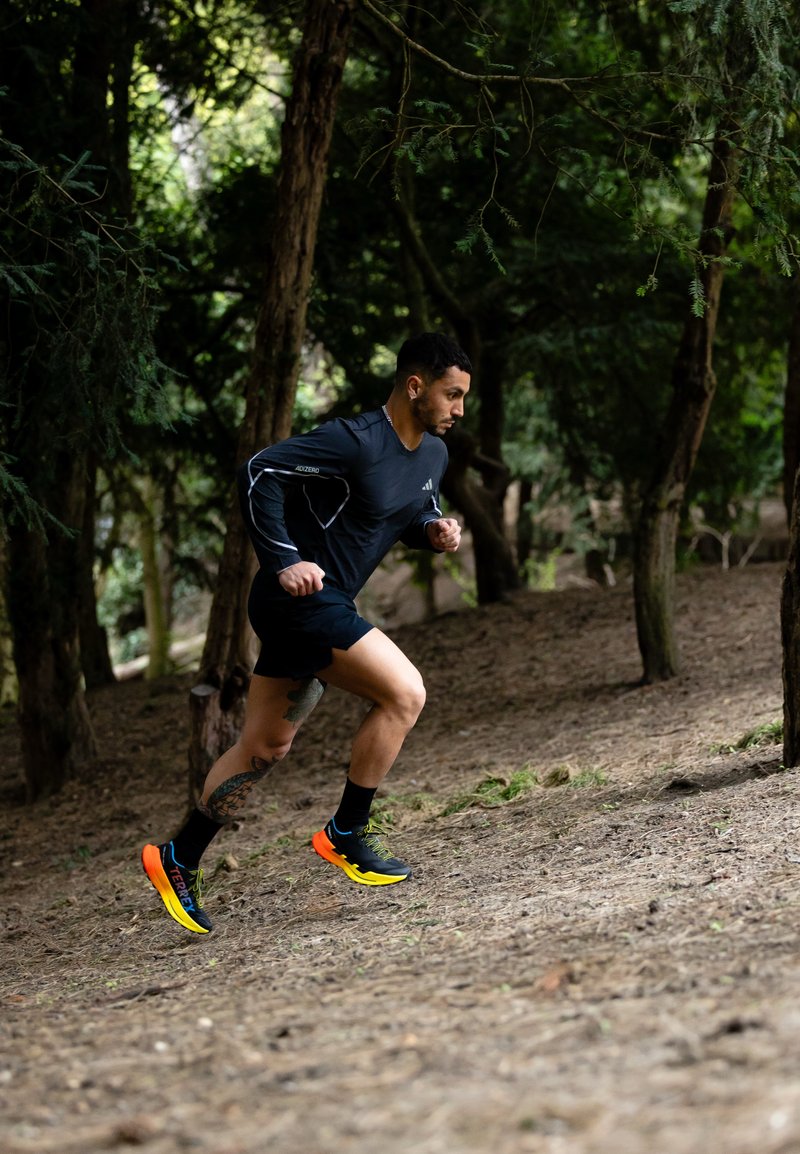 Man in zwarte sportkleding rent heuvelop op een onverharde route omgeven door hoge groene bomen in een bos.