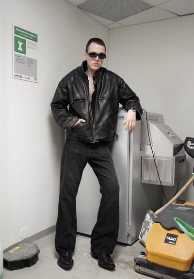 Chaqueta de cuero negra con cremallera frontal, camiseta oscura debajo y jeans negros holgados. Gafas de sol oscuras y zapatos negros. Fondo industrial.