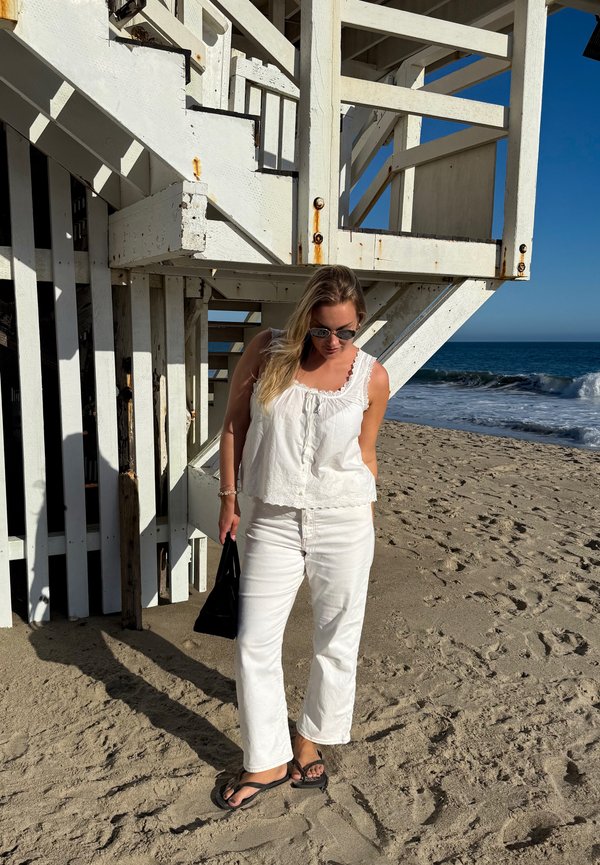 Žena v bielom topu a nohaviciach so slnečnými okuliarmi stojí na piesočnatej pláži pri veži záchranárov s oceánskymi vlnami v pozadí.