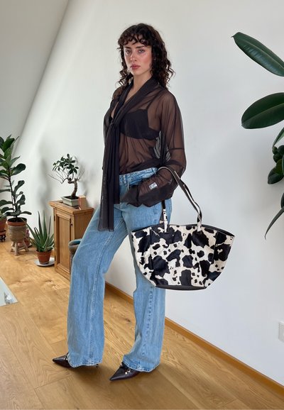 Blusa negra transparente con detalle de lazo, jeans de mezclilla oscuros acampanados y bolso de estampado de vaca. Tacones marrones de punta, plantas de interior en el fondo.