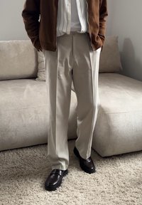 Personne portant un pantalon beige, une chemise blanche, une veste marron et des chaussures marron foncé, debout sur un sol tapis près d'un canapé beige.