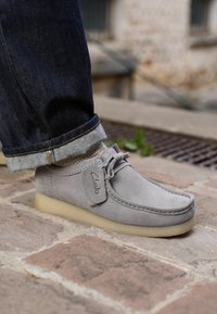 Zapato Clarks de ante gris con suela de crepé, usado con calcetines beige y vaqueros oscuros remangados sobre pavimento de piedra.