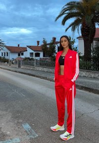 Fato de treino vermelho da Adidas com riscas brancas nas mangas e laterais, combinado com um top curto preto e ténis multicoloridos, em pé numa rua.