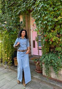 Τζιν πουκάμισο με τσέπες, ανοιχτό μπλε μακριά φούστα με μπροστινή σχισμή, καφέ ζώνη και καφέ μποτάκια, σε φόντο πράσινης κισσού και ροζ πόρτας.