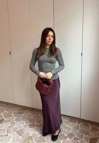 Langer burgunderfarbener Satinrock kombiniert mit einem taillierten grauen Langarmshirt. Halte eine strukturierte burgunderfarbene Handtasche. Neutralfarbener Steinboden im Hintergrund.