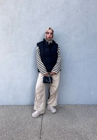 Μαύρο πλούσιο γιλέκο, ριγέ μακρυμάνικο πουκάμισο, μπεζ παντελόνι με φαρδιές γραμμές, ουδέτερα παπούτσια και μια μικρή μαύρη τσάντα με αλυσίδα πάνω σε μια υφασμάτινη επιφάνεια.