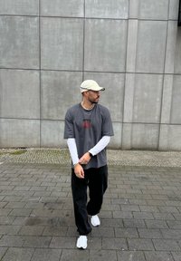 Γκρι φαρδύ t-shirt με κόκκινη γραφική παράσταση, σε στρώσεις πάνω από ένα λευκό που�κάμισο με μακριά μανίκια. Μαύρα παντελόνια και λευκά αθλητικά παπούτσια, με ένα ανοιχτόχρωμο καπέλο.