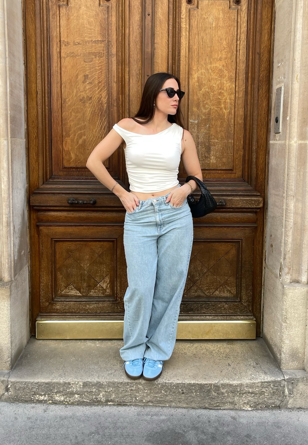 Λευκή μπλούζα χωρίς ώμους, ανοιχτό μπλε παντελόνι φάρσας και μπλε αθλητικά παπούτσια, στέκεται απέναντι σε μια καφέ ξύλινη πόρτα με περίπλοκες λεπτομέρειες.