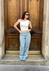 Λευκή μπλούζα χωρίς ώμους, ανοιχτό μπλε παντελόνι φάρσας και μπλε αθλητικά παπούτσια, στέκεται απέναντι σε μια καφέ ξύλινη πόρτα με περίπλοκες λεπτομέρειες.