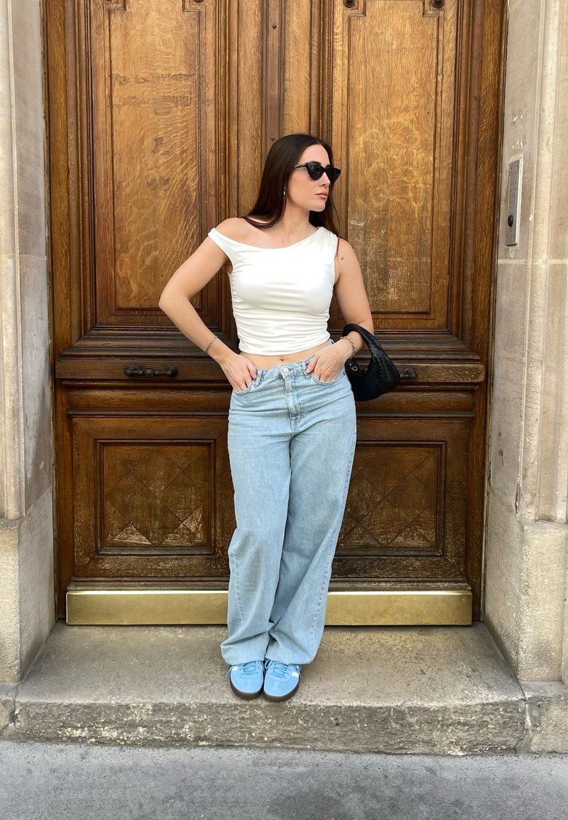 Λευκή μπλούζα χωρίς ώμους, ανοιχτό μπλε παντελόνι φάρσας και μπλε αθλητικά παπούτσια, στέκεται απέναντι σε μια καφέ ξύλινη πόρτα με περίπλοκες λεπτομέρειες.