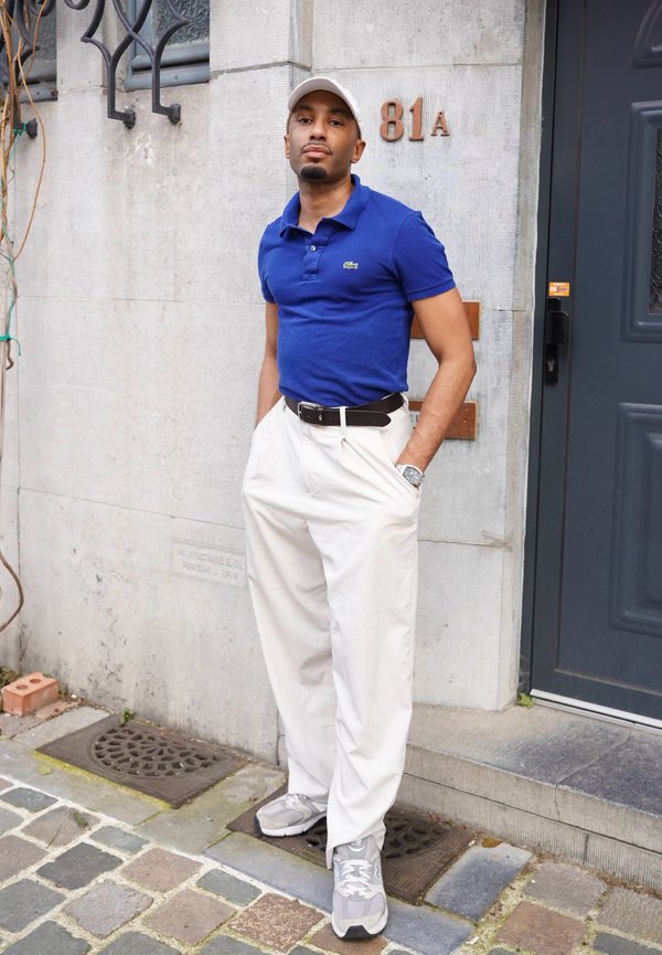 Mann in blauem Poloshirt, weissen Hosen und grauen Sneakers steht mit den Händen in den Taschen beim grauen Eingang eines Gebäudes mit der Nummer 81A.