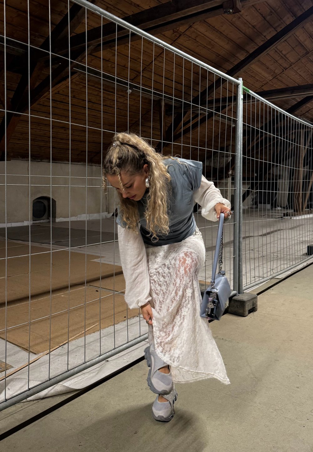 Žena upravuje bielu čipkovanú sukňu, zatiaľ čo má na sebe šedé tenisky a drží zodpovedajúcu šedú kabelku. Stojí pri kovovom plote v podkrovnom priestore.