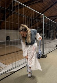 Žena upravuje bielu čipkovanú sukňu, zatiaľ čo má na sebe šedé tenisky a drží zodpovedajúcu šedú kabelku. Stojí pri kovovom plote v podkrovnom priestore.