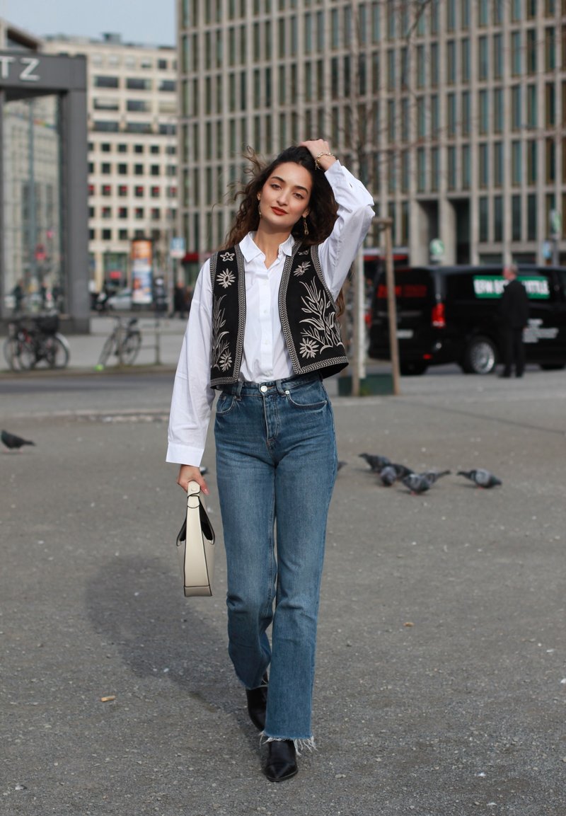 Μαύρο κεντημένο γιλέκο πάνω από λευκό πουκάμισο, μπλε χαμηλόμεσα τζιν και μαύρες μπότες μέχρι τον αστράγαλο. Κρατώντας μια κρεμ τσάντα. Αστικό φόντο.