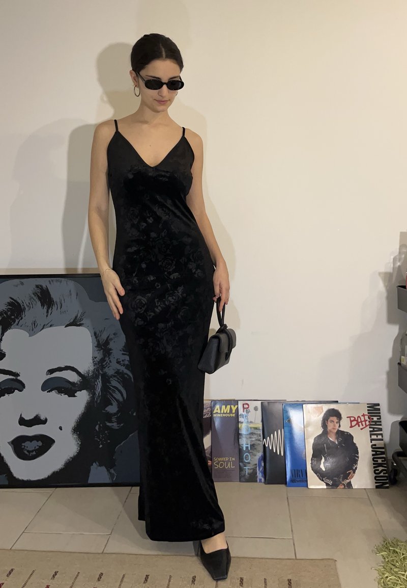 Femme portant une longue robe noire et des lunettes de soleil, debout à l'intérieur, tenant un petit sac à main noir, près d'une œuvre encadrée de Marilyn Monroe et de disques vinyles.