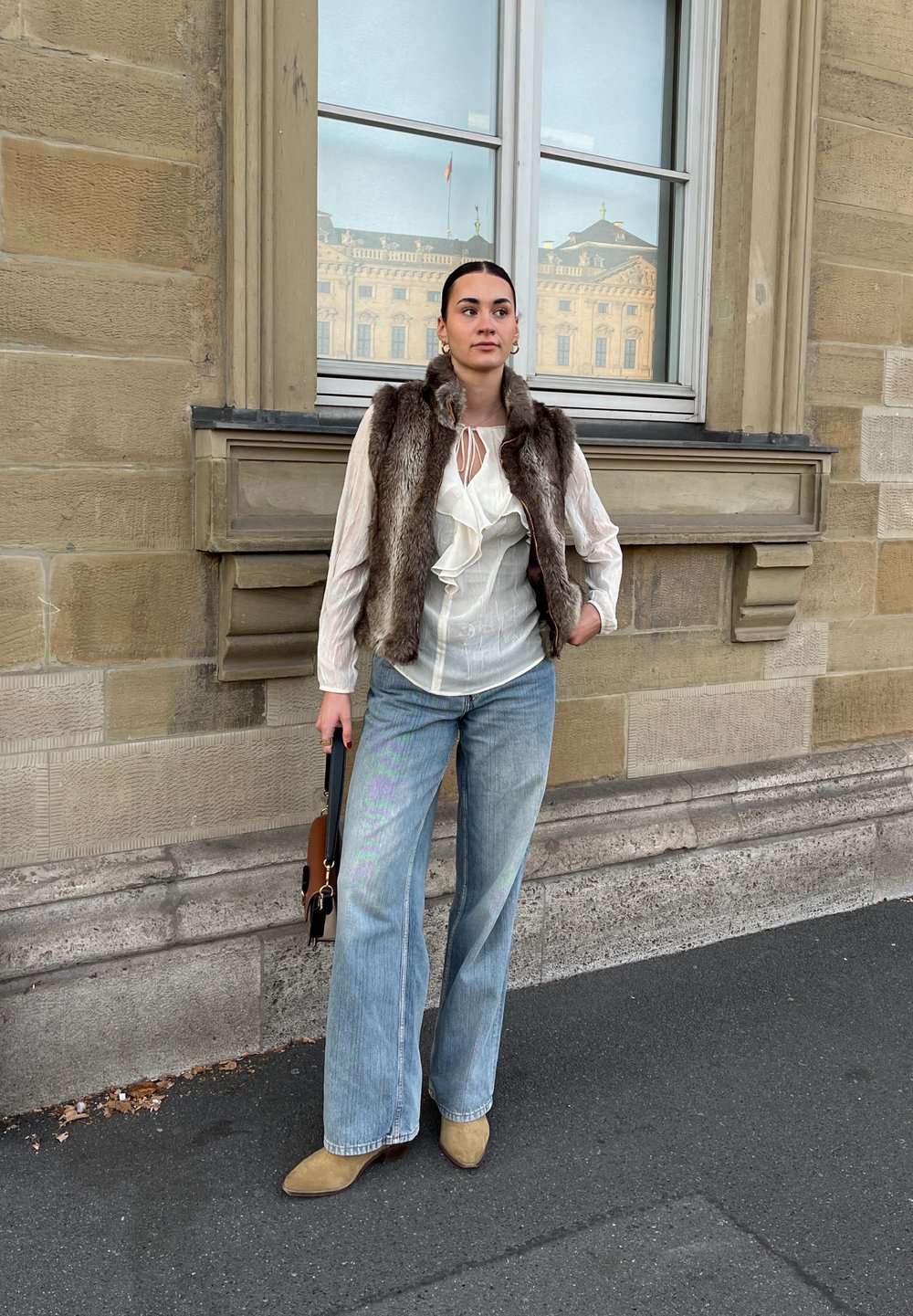 Colete de pelúcia, blusa branca com babados, jeans de denim claro de perna larga e botins bege. Os acessórios incluem uma pequena mala castanha.