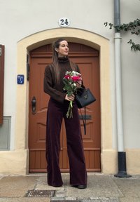 Brown ribbed turtleneck jumper, wide-leg burgundy trousers, holding a bouquet of pink roses, black handbag, standing by a brown door with the number 24.