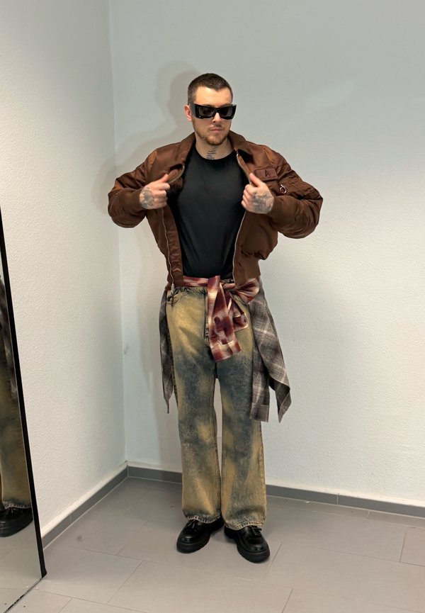 Un homme portant des lunettes de soleil, une veste brune, une chemise noire et un jean ample pose dans une cabine d’essayage en tenant sa veste ouverte.