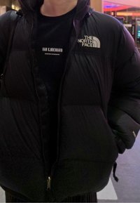 Čierna nadýchaná bunda na zips vpredu, s logom The North Face na pravom hornom boku. Nosená cez čierne tričko s bielym textom.