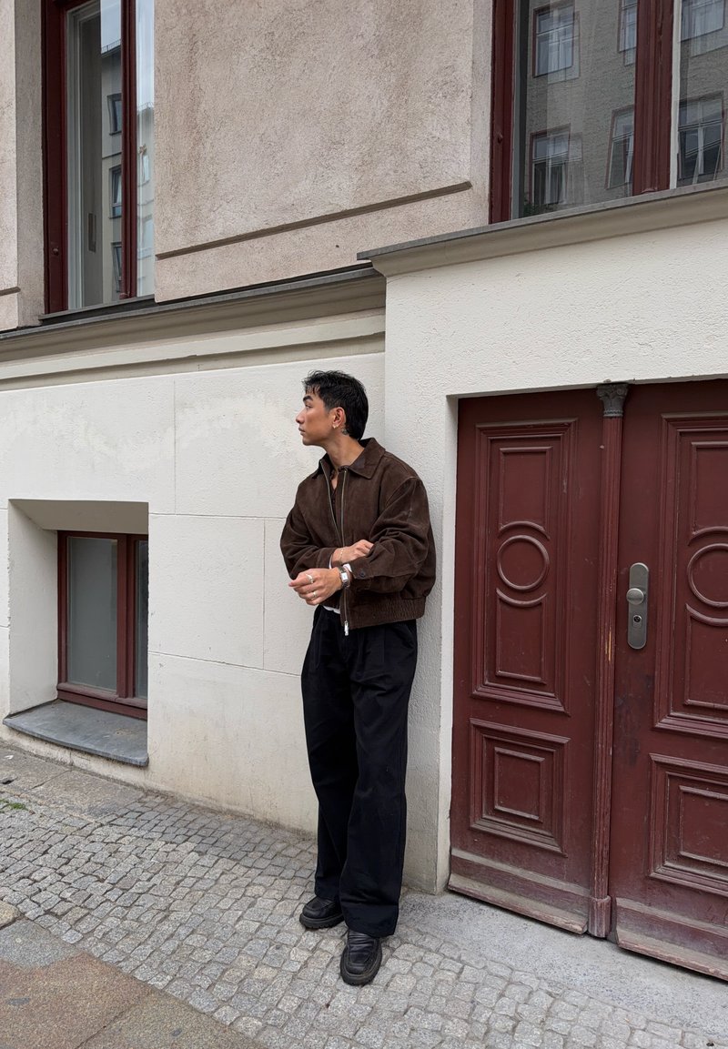 Homme portant une veste marron et un pantalon noir se tient dans une rue pavée à côté d'un mur blanc et d'une porte rouge foncé, regardant vers sa gauche.