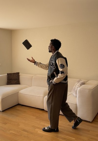 Hombre de pie en una habitación beige, lanzando una bolsa negra con la mano derecha, vistiendo una chaqueta universitaria negra y blanca y pantalones marrones cerca de un sofá beige.