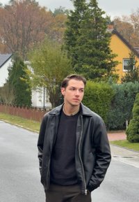 Zwarte leren jas over een donkere trui, staande op een straat met groenvoorzieningen en woonhuizen op de achtergrond. Bewolkte lucht.