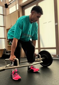 Atleta w tealowej koszulce Adidas podnosi srebrny hex bar, nosząc czarne spodnie i jaskraworóżowe buty sportowe, na podłodze siłowni.