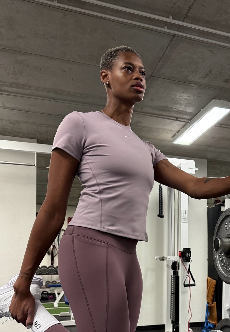 Femme en tenue de sport mauve étirant son muscle quadriceps dans une salle de sport, tenant son pied derrière elle avec une main, l'autre main reposant sur une machine de musculation.