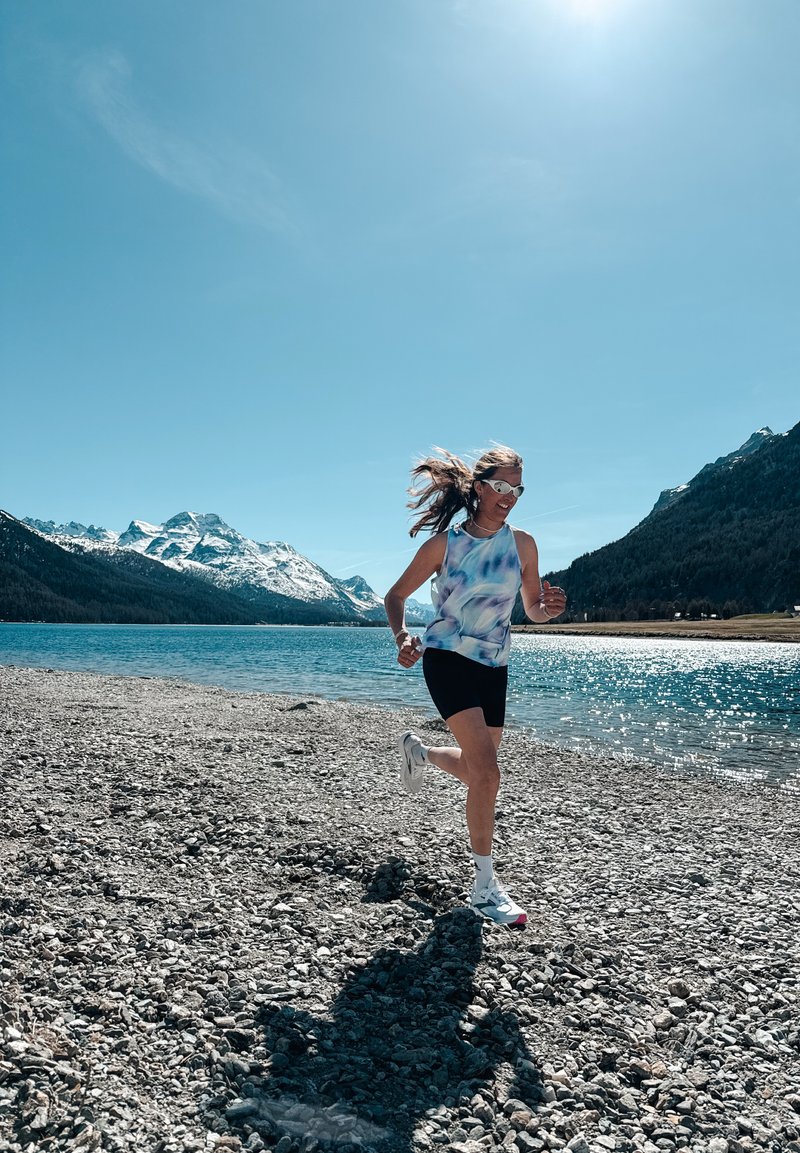 Femme courant sur un rivage rocheux d'un lac avec des montagnes enneigées et un ciel bleu clair en arrière-plan lors d'une journée ensoleillée.