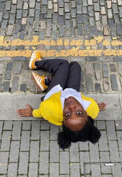 Persona sentada sobre los adoquines, vistiendo un cárdigan amarillo sobre una camisa blanca, pantalones a rayas y zapatillas naranjas. Líneas amarillas visibles cerca.