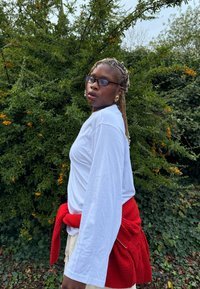 Hosszú ujjú fehér ing piros pulóverrel, amelyet derékvonalban kötöttek össze; fonott haj és szemüveg egészíti ki a megjelenést a zöld növényzet előtt.