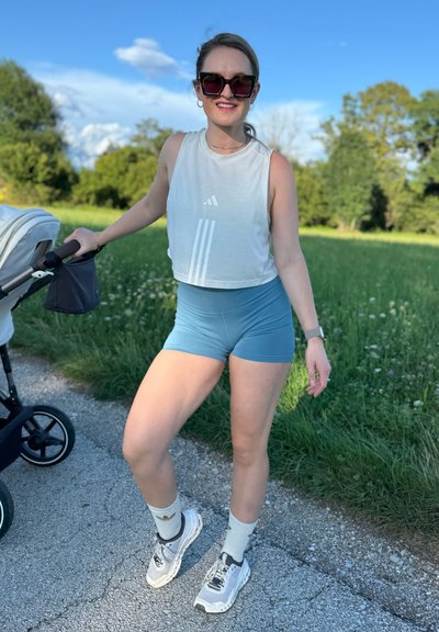 Top sin mangas de color gris claro con tres franjas blancas, pantalones cortos azules, zapatillas blancas con detalles grises y calcetines de estampado, de pie cerca de un carrito.