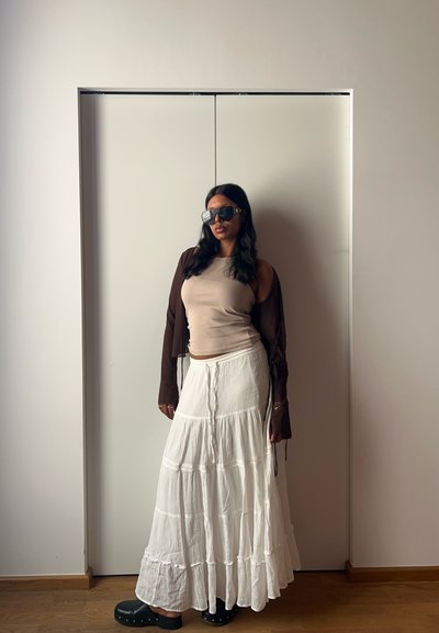 Camisa marrón de manga larga, top de tirantes beige y una falda maxi blanca de volantes. Zuecos negros con acentos de tachuelas. Gafas de sol completan el look.