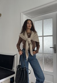 Brun ribstrikket langærmet top, cremefarvet striktrøje bundet ved halsen, blå højtaljede jeans, sort bucket-taske, hvid dør med tekstur i baggrunden.