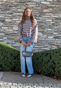 Grijze gestreepte trui met brede mouwen en hoge kraag; blauwe high-waisted jeans; zilveren spitse hakken; kleine zwarte handtas; steenachtige muur op de achtergrond.