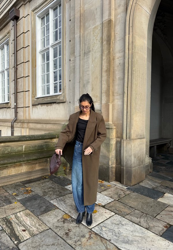 Une femme en long manteau brun tient un petit sac à main devant un bâtiment en pierre aux hautes fenêtres et à l’entrée voûtée.