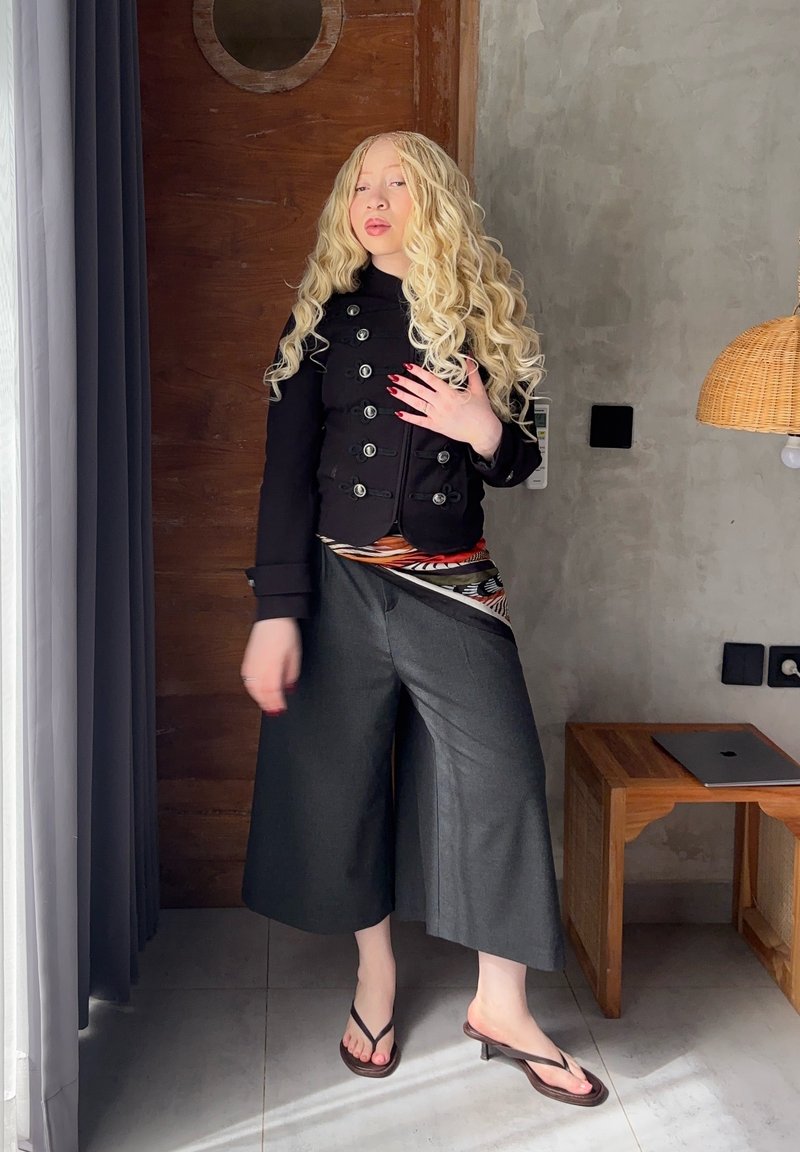 Femeie blondă cu păr lung și creț, purtând o jachetă neagră, pantaloni largi gri și sandale, stă în interior lângă o ușă de lemn și o masă cu laptop.