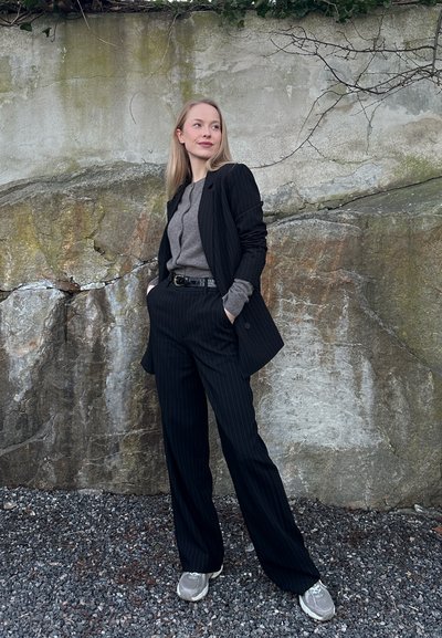 Mujer con traje de rayas y suéter gris de pie sobre grava frente a una pared de roca texturizada con ramas desnudas arriba.