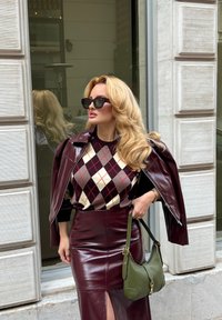 Donna con capelli biondi che indossa occhiali da sole neri, maglione a rombi, giacca in pelle bordeaux drappeggiata sulle spalle, gonna abbinata e tiene una borsa verde.