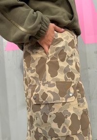 Bež kamuflažne cargo hlače s velikim bočnim džepovima, s sjajnim dugmadima za kopčanje i opuštenim krojem. Vidljiva teksturirana tkanina.