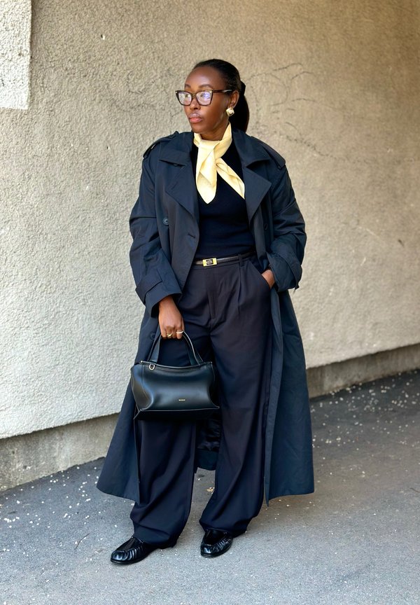 Frau in schwarzem Trenchcoat, weit geschnittenen schwarzen Hosen, gelbem Schal und Brille, die eine schwarze Handtasche hält und mit der Hand in der Tasche vor einer strukturierten Wand steht.