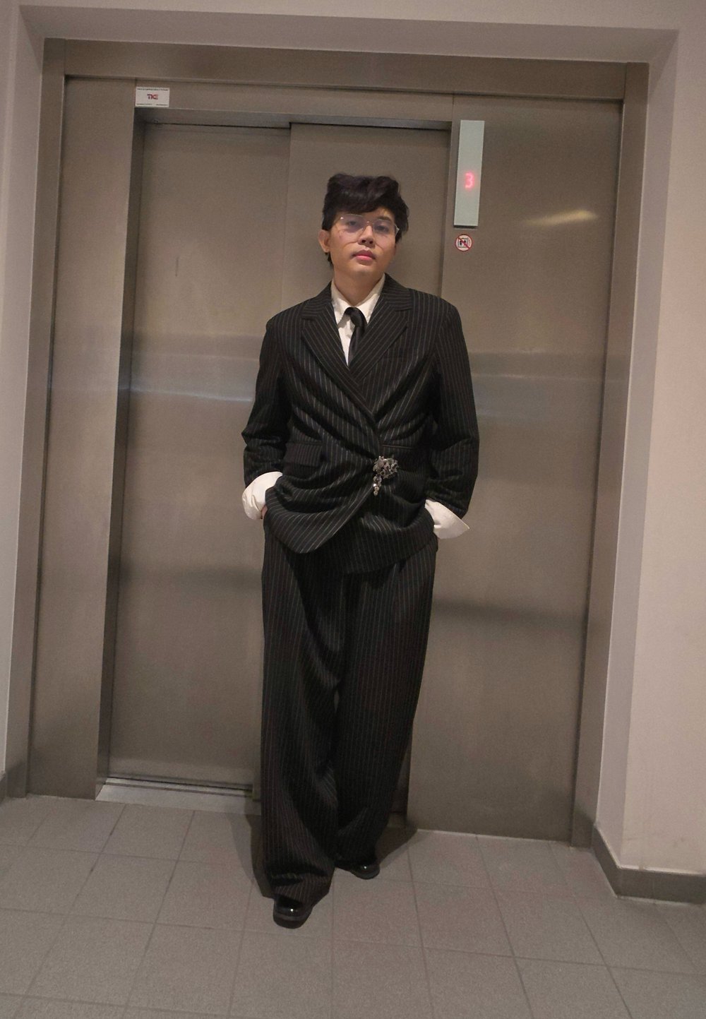 Costume à rayures noires avec une veste cintrée et un pantalon large, assorti d'une chemise blanche et d'une cravate noire. Une broche remarquable est placée à la taille.