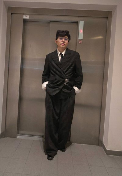 Traje de rayas negras con una chaqueta entallada y pantalones anchos, combinado con una camisa blanca y una corbata negra. Un broche notable en la cintura.