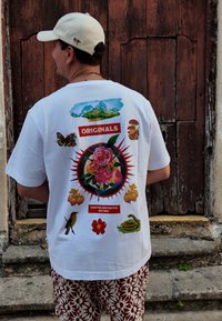 Άνδρας που φοράει λευκό καπέλο και λευκό μπλουζάκι με πολύχρωμα γραφικά με λουλούδια, ζώα και φύση, στέκεται δίπλα σε μια φθαρμένη ξύλινη πόρτα.