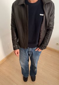 Άτομο που φοράει σκούρο καφέ δερμάτινο μπουφάν, μπλε πουλόβερ με ριμπ ύφανση και ετικέτα "GUESS JEANS", άνετα μπλε τζιν και καφέ παπούτσια, στέκεται σε εσωτερικό χώρο.