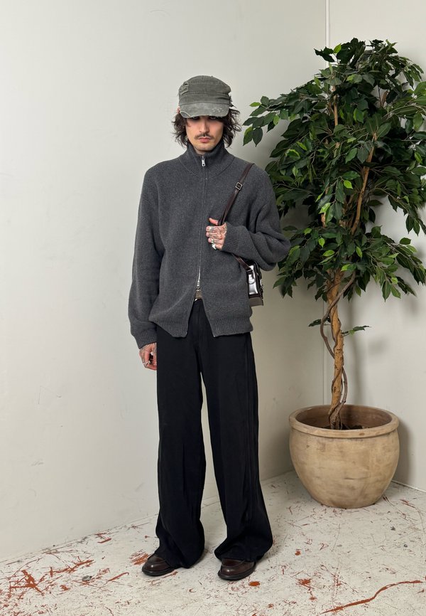 Person, die einen grauen Zip-up-Pullover, schwarze weit geschnittene Hosen, eine olivgrüne Mütze trägt und eine braune Schultertasche bei sich hat, steht neben einer großen Topfpflanze.