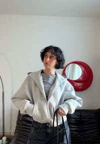 Sweat à capuche gris clair avec fermeture éclair, doté d'une grande capuche, porté avec un t-shirt rayé noir et blanc et un jean foncé. Sac noir à la main. Miroir rond rouge en arrière-plan.